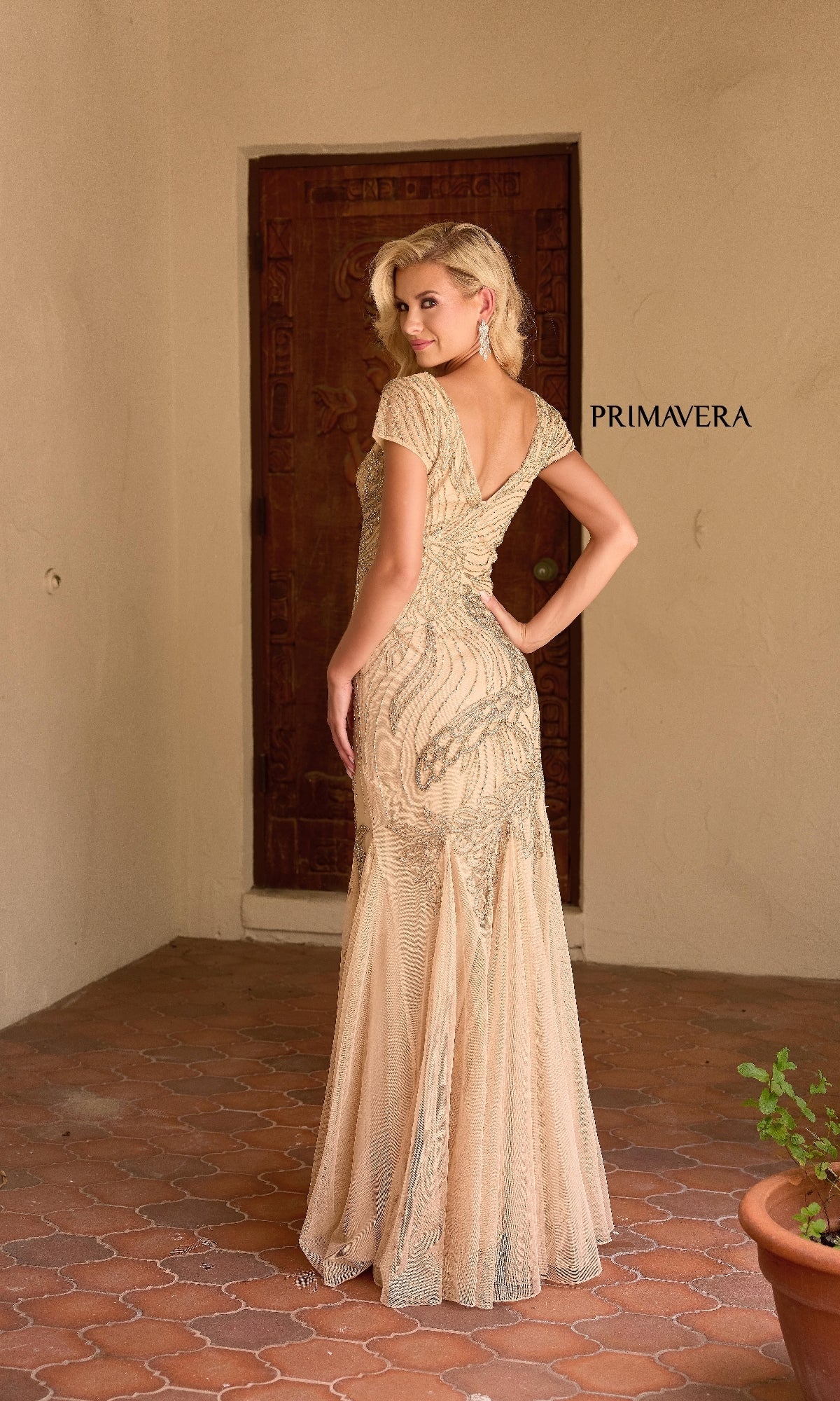 A woman poses with her back to the camera in front of a wooden door, wearing the Primavera 13148 long beige formal dress with fit-and-flare silhouette and elegant beading.